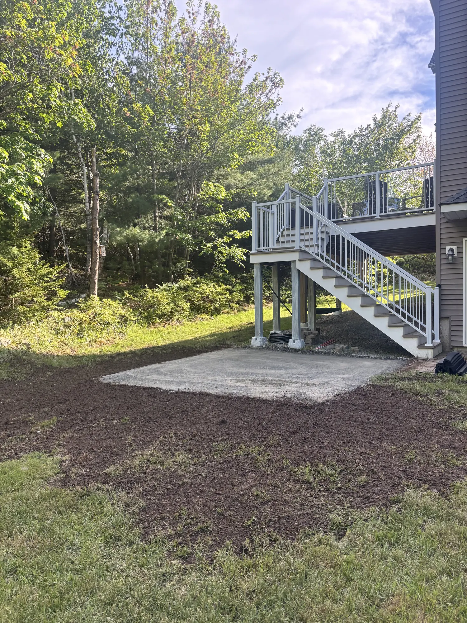 Freshly graded backyard soil and a concrete pad below an elevated deck with white stairs