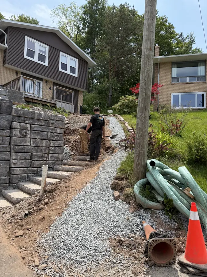 Stone retaining wall steps beside a gravel drainage trench with corrugated pipe and a crew member working — hardscaping project in Halifax, Nova Scotia